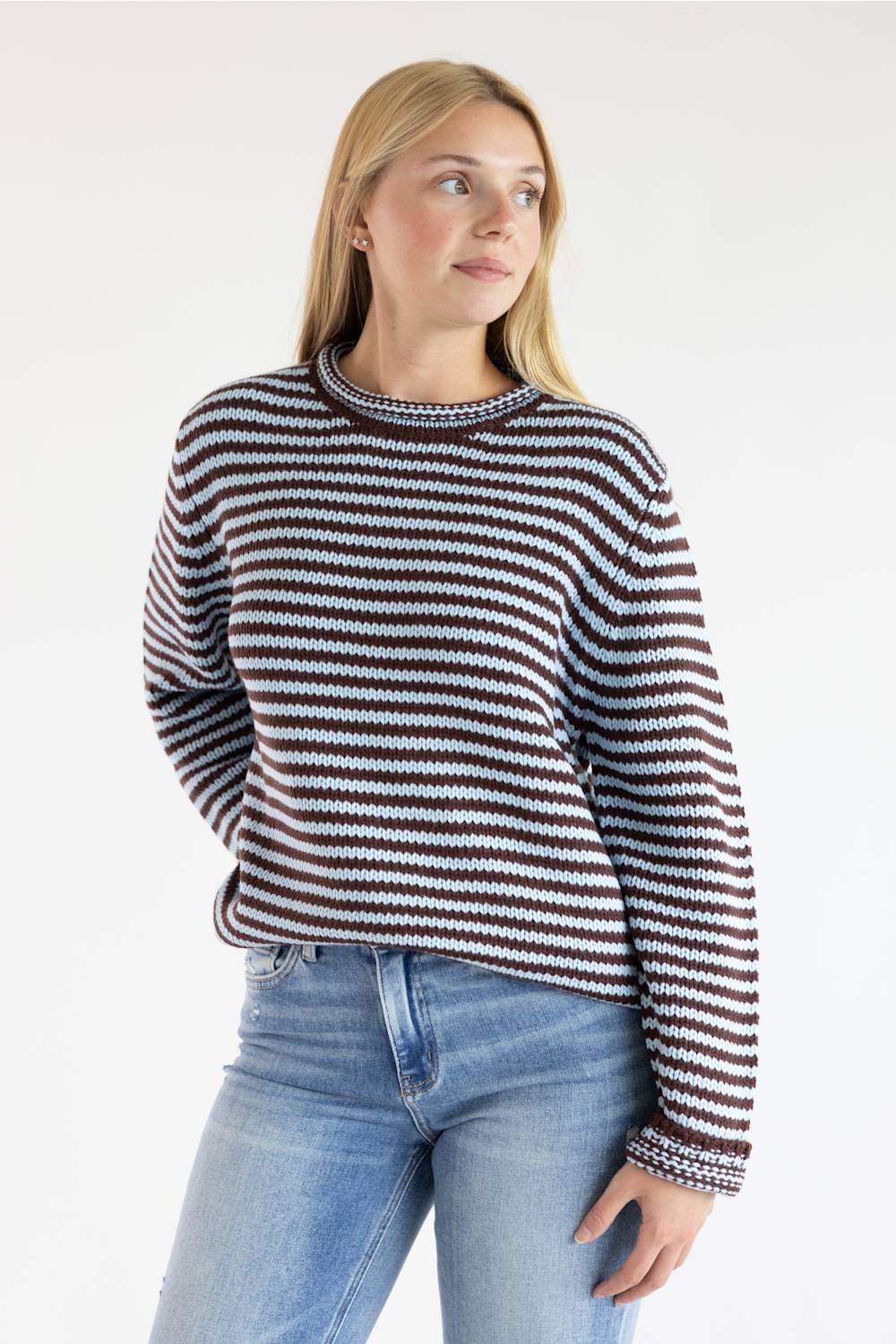 A woman wearing a long-sleeve knit sweater with horizontal brown, black, and light blue stripes, paired with light-wash blue jeans, against a plain white studio background.