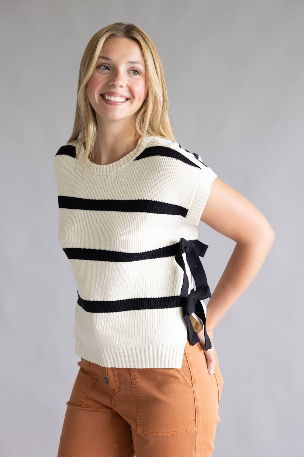 A smiling blonde woman wearing a cream ribbed knit sweater vest with black horizontal stripes and a side bow detail, paired with brown pants, against a neutral gray studio background.