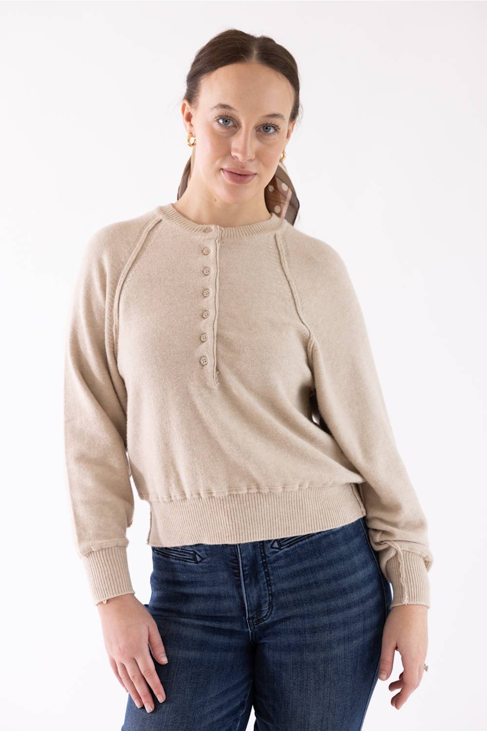 A woman wearing a light beige knit sweater with a buttoned henley-style neckline and dark blue jeans, standing against a plain white studio background.