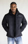 A man wearing a black Ariat quilted hooded jacket over a black shirt and blue jeans, standing against a plain light gray background.