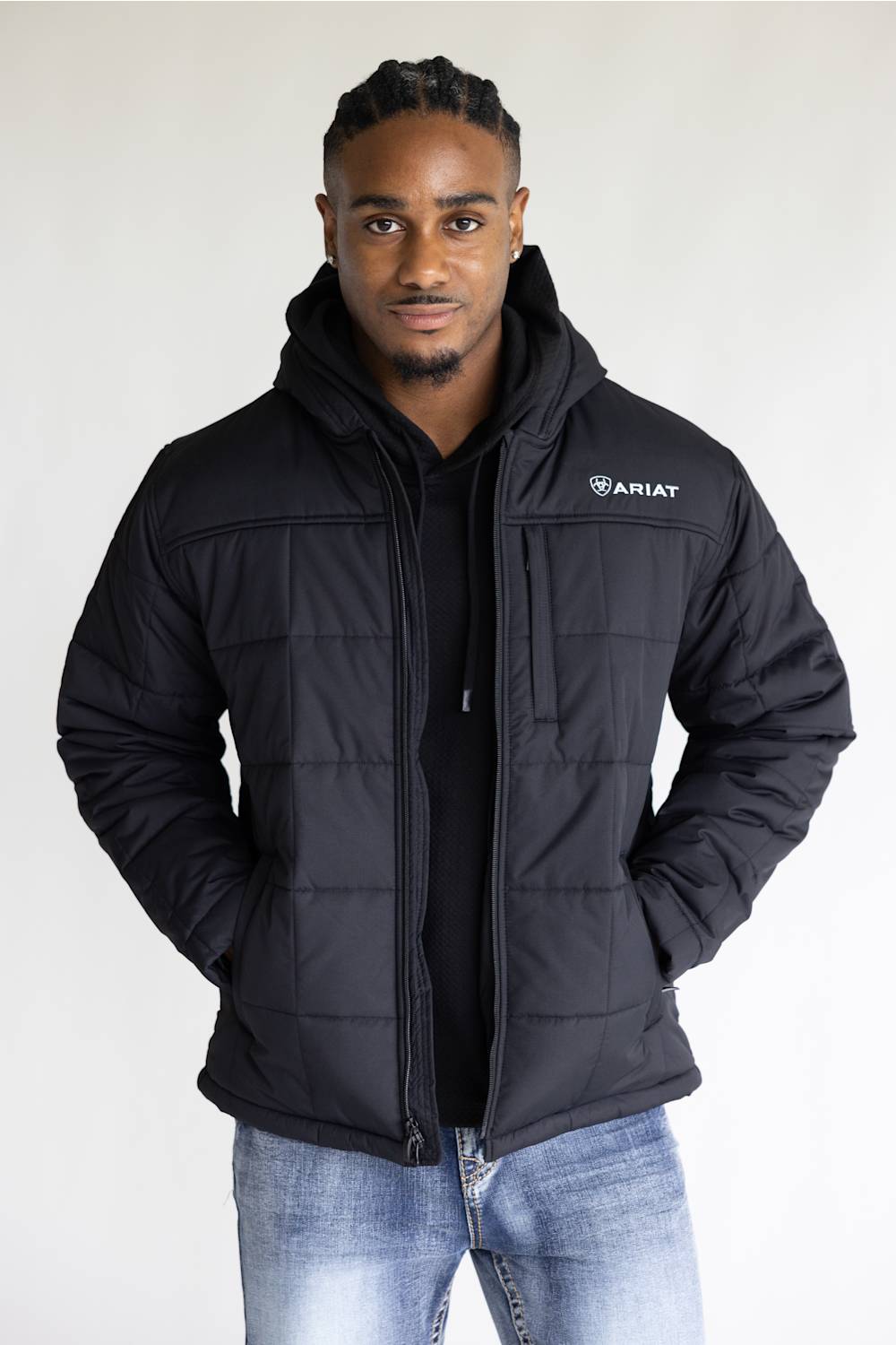 A man wearing a black Ariat quilted hooded jacket over a black shirt and blue jeans, standing against a plain light gray background.