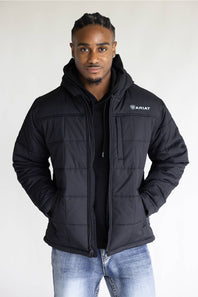 A man wearing a black Ariat quilted hooded jacket over a black shirt and blue jeans, standing against a plain light gray background.