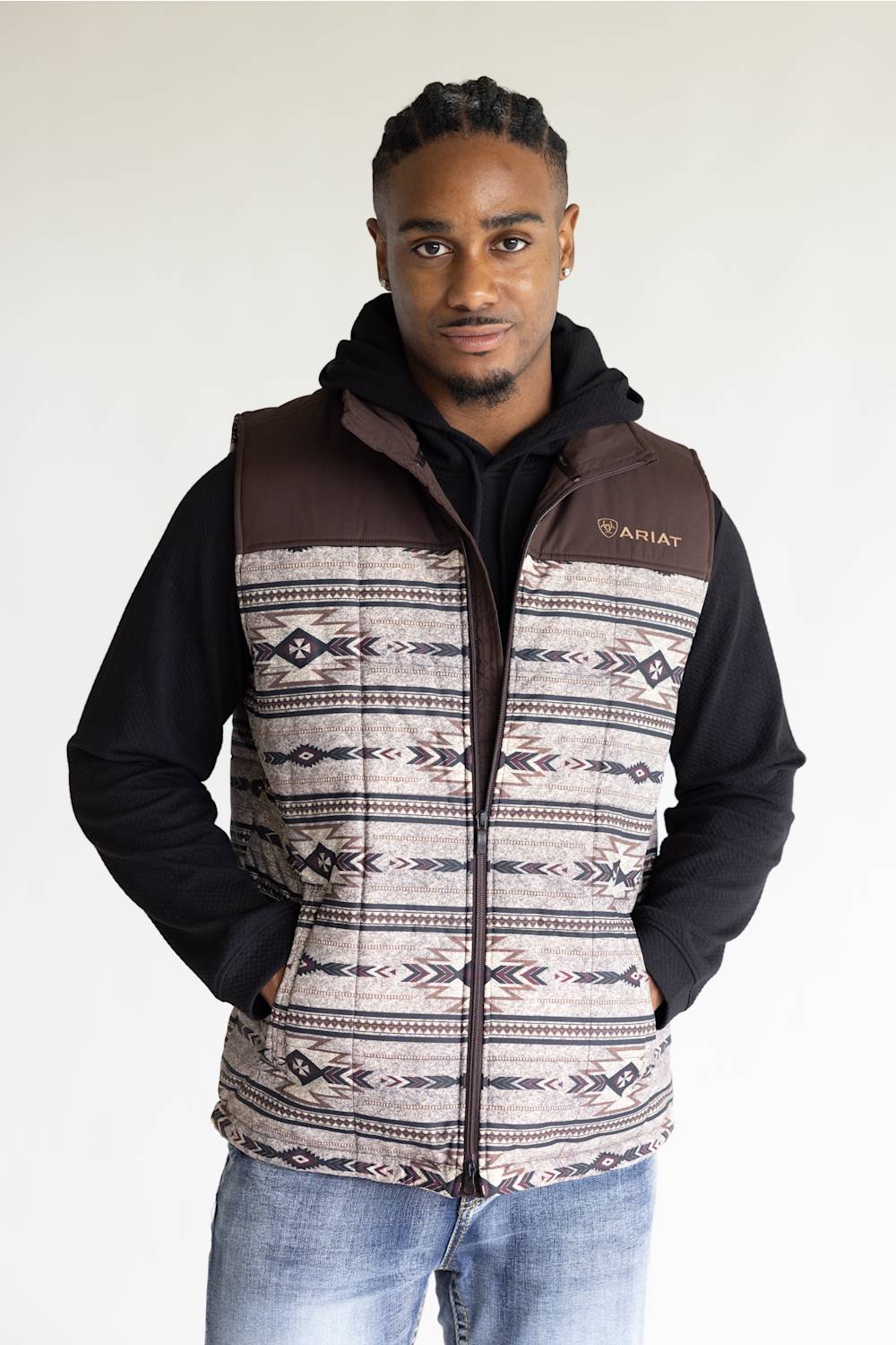 A man wearing a brown and beige patterned zip-up vest over a black hoodie and blue jeans, standing against a plain light background. The vest features a geometric Southwestern-style pattern and a small 'Ariat' logo on the left chest.
