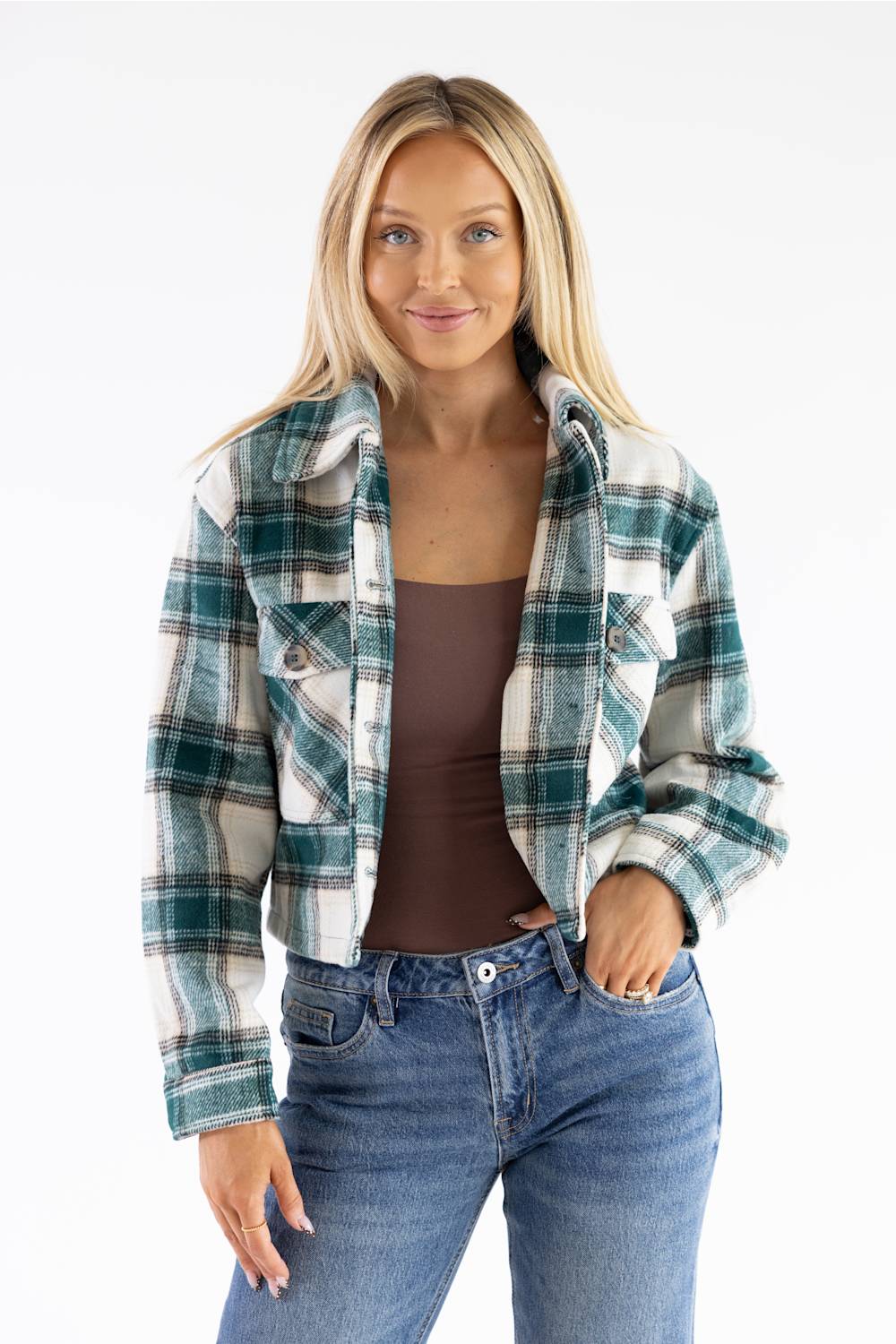 A woman modeling a cropped, long-sleeve plaid flannel shirt jacket in green, white, and blue, worn open over a brown tank top and blue jeans against a plain white background.