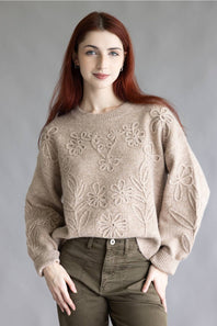 A woman with long red hair wearing a beige knit sweater with raised floral patterns and olive green pants, standing against a plain gray studio background.