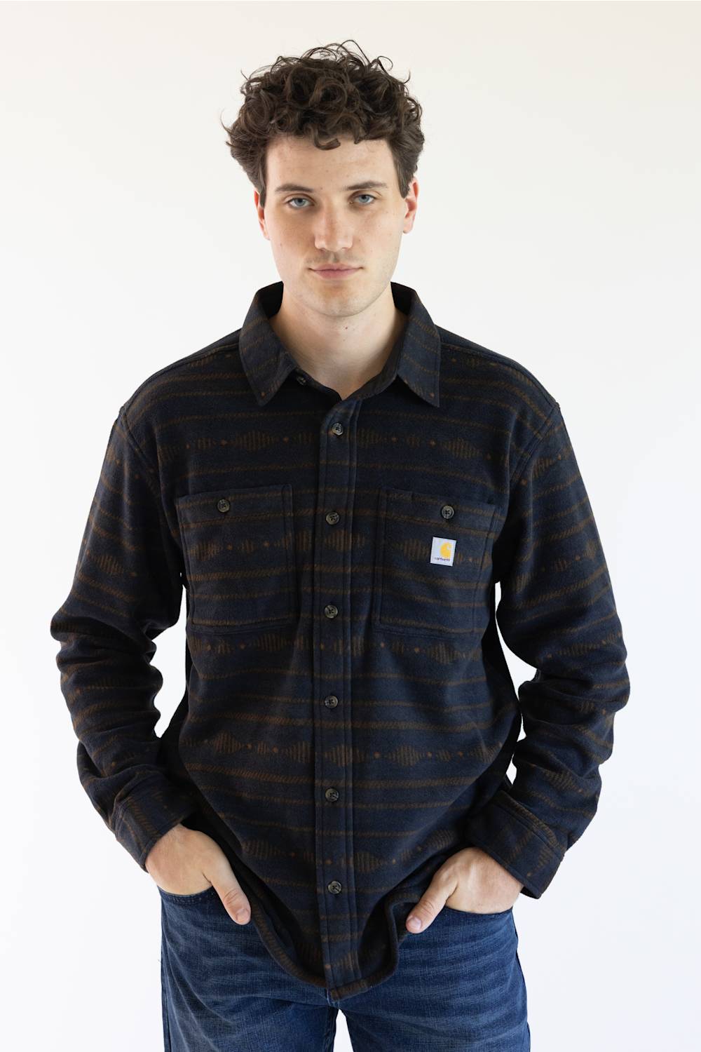 A man wearing a long-sleeve Carhartt plaid button-up shirt in navy and brown with thin orange stripes, paired with blue jeans, against a plain white background.