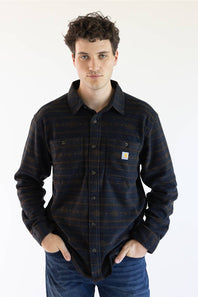 A man wearing a long-sleeve Carhartt plaid button-up shirt in navy and brown with thin orange stripes, paired with blue jeans, against a plain white background.