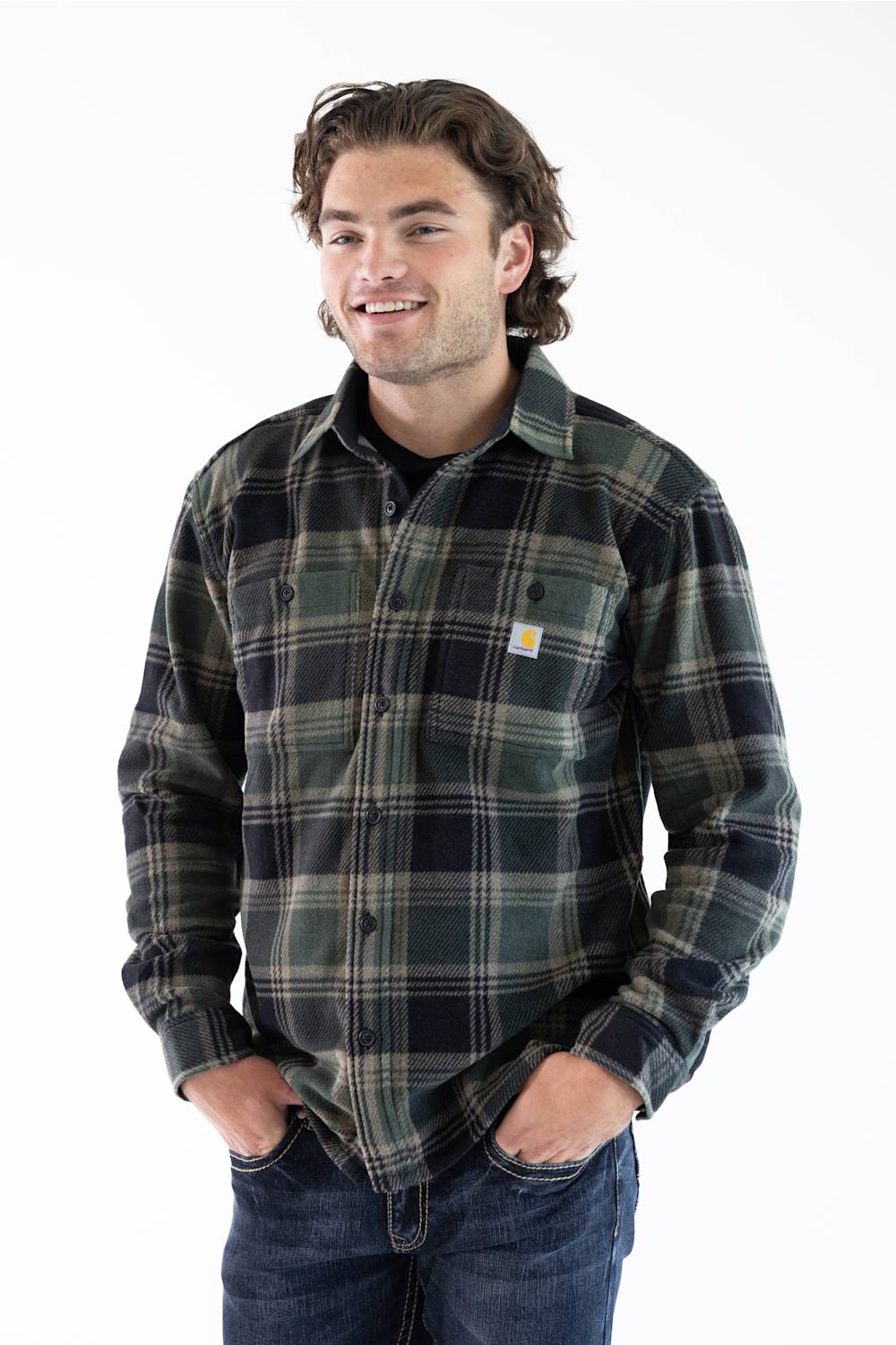 A man wearing a long-sleeve plaid button-up shirt in dark green, black, and gray, paired with blue jeans, standing against a plain white background.