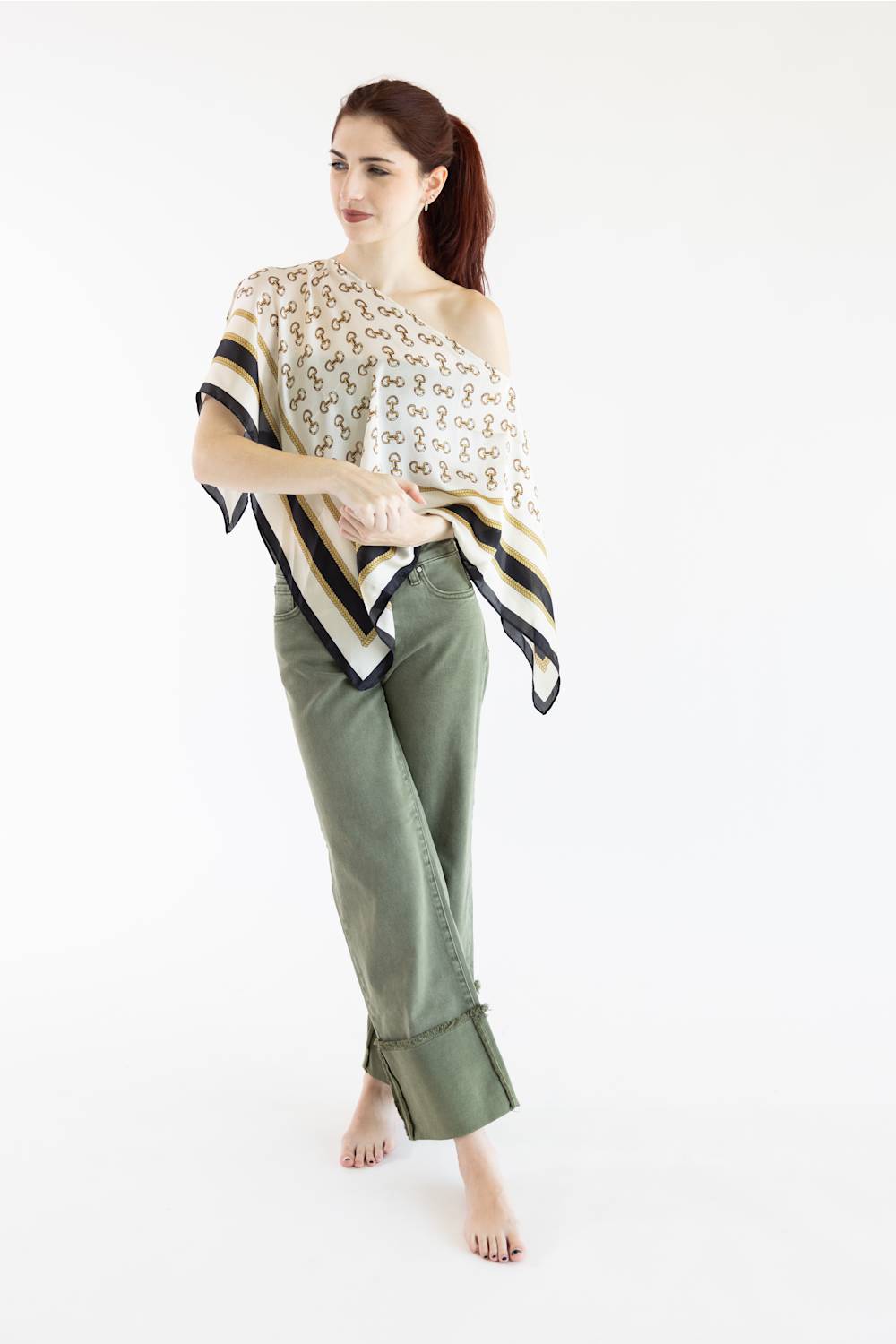 A woman modeling a one-shoulder draped top with a beige, black, and white patterned design, paired with olive green pants, against a plain white studio background.