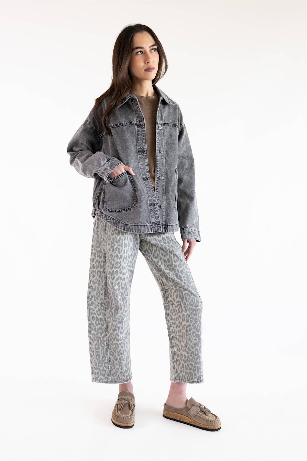 A model stands against a plain white studio background wearing a washed gray denim jacket, gray leopard-print pants, a beige top, and brown slip-on shoes.