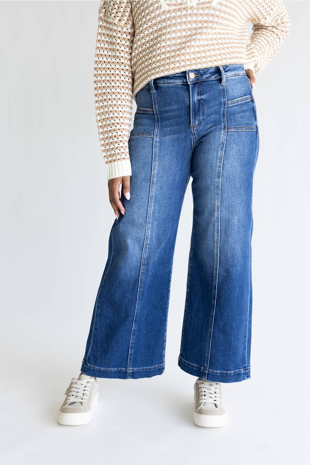 A person wearing blue wide-leg denim jeans with a cream and brown patterned knit sweater and white sneakers, shown from the chest down against a light gray studio background.