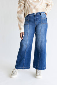 A person wearing blue wide-leg denim jeans with a cream and brown patterned knit sweater and white sneakers, shown from the chest down against a light gray studio background.