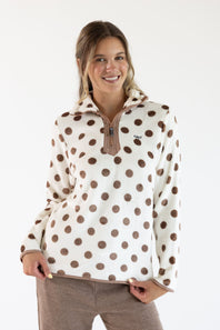 A smiling woman wearing a cream fleece pullover with brown polka dots and a brown zipper, paired with brown corduroy pants, against a plain white studio background.