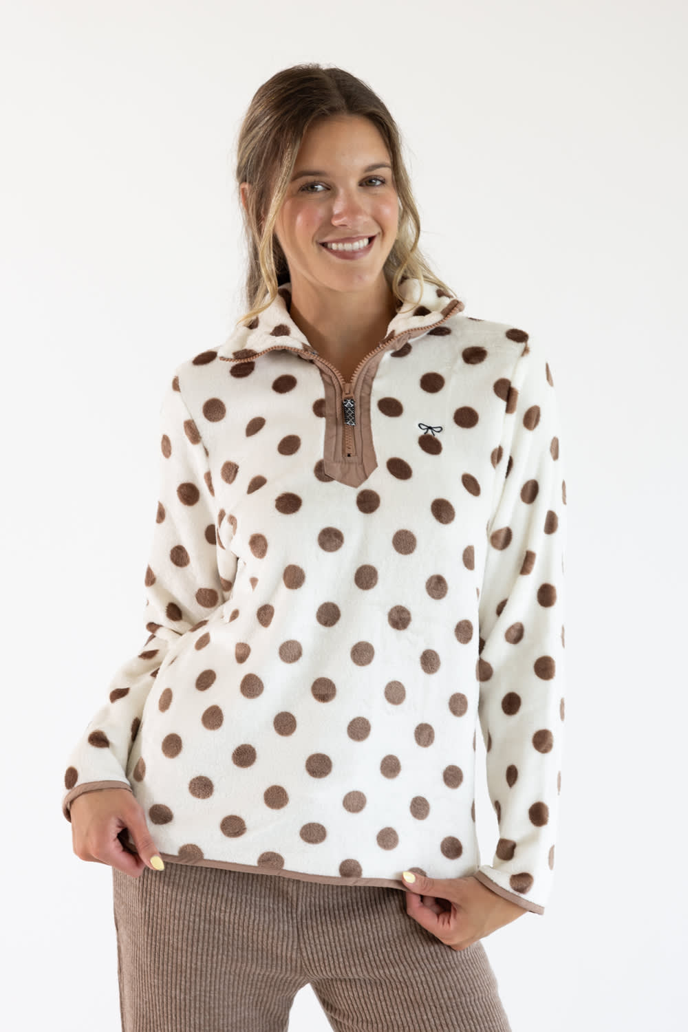 A smiling woman wearing a cream fleece pullover with brown polka dots and a brown zipper, paired with brown corduroy pants, against a plain white studio background.