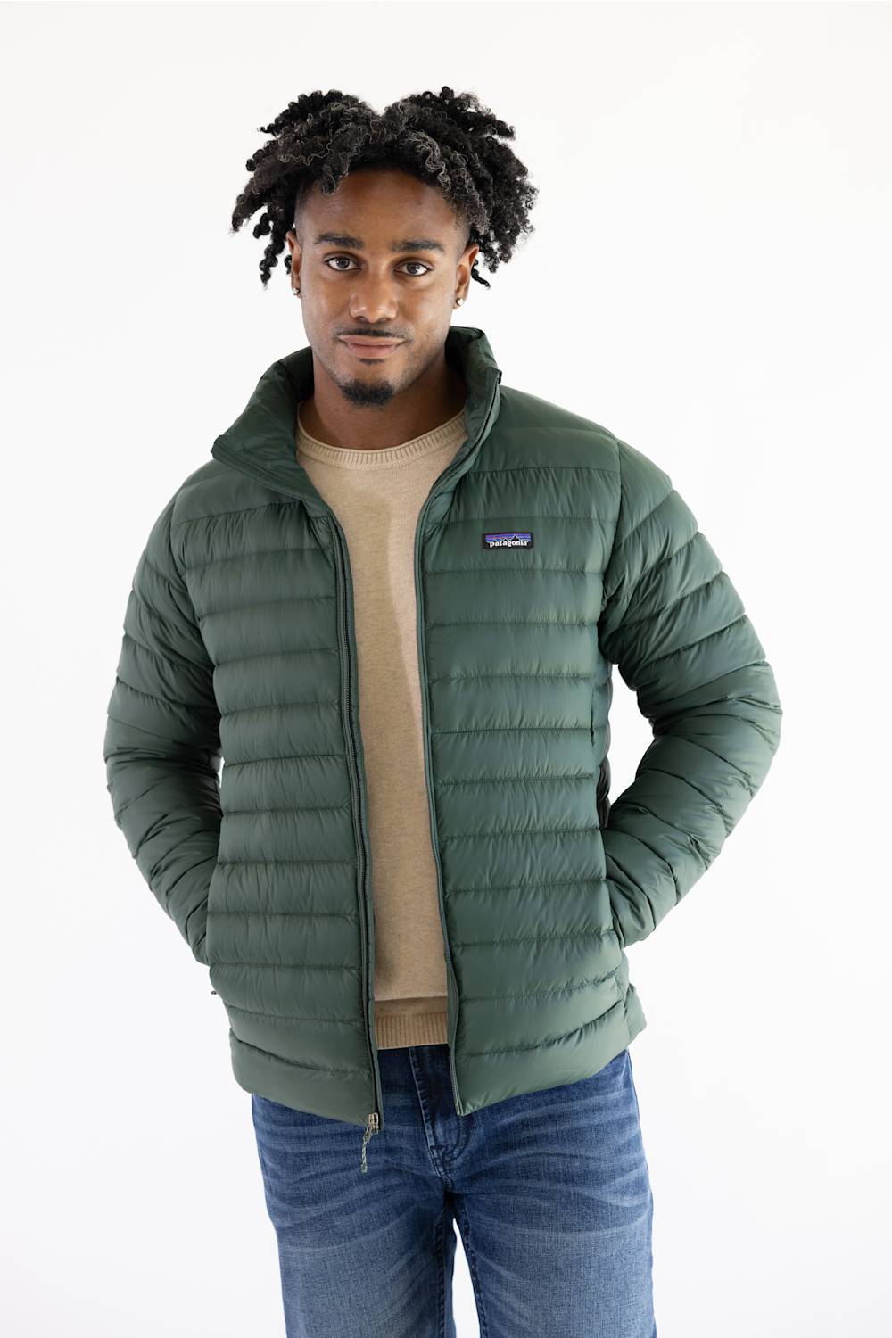 A man wearing a green Patagonia puffer jacket over a beige t-shirt and blue jeans, standing against a plain white background.