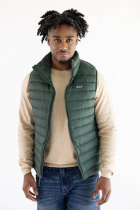 A man wearing a green Patagonia puffer vest over a beige sweater and blue jeans, standing against a plain white background.