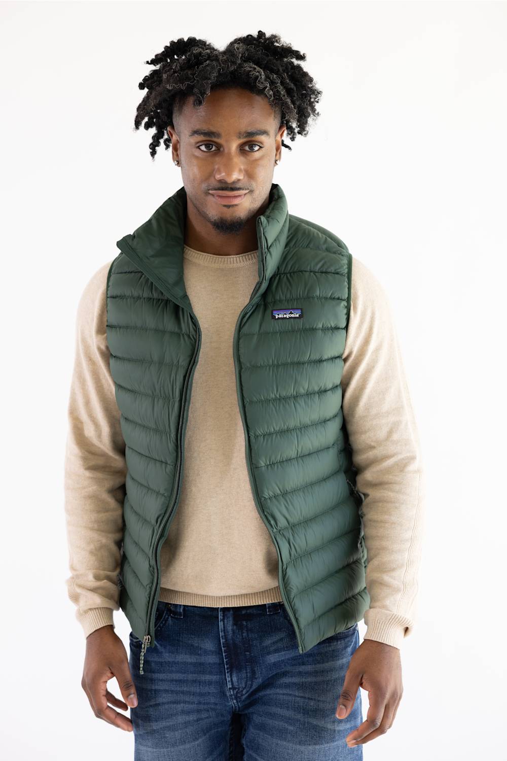 A man wearing a green Patagonia puffer vest over a beige sweater and blue jeans, standing against a plain white background.