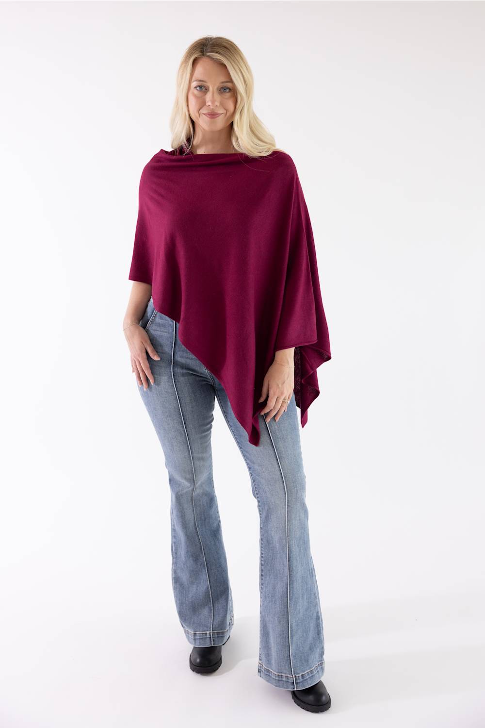 A woman modeling a draped, deep burgundy poncho-style top with light-wash blue jeans and black shoes, standing against a plain white studio background.
