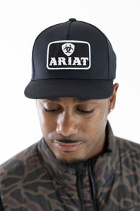 Front view of a man wearing a black baseball cap featuring a white embroidered patch on the front with Ariat's logo and the word 'ARIAT'. He is wearing a camouflage-patterned jacket, against a plain white background.