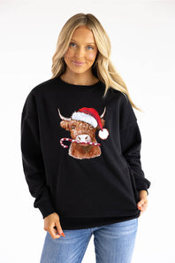 A woman wearing a black crewneck sweatshirt with a graphic of a Highland cow wearing a red Santa hat and holding a candy cane, paired with blue jeans, against a plain white background.
