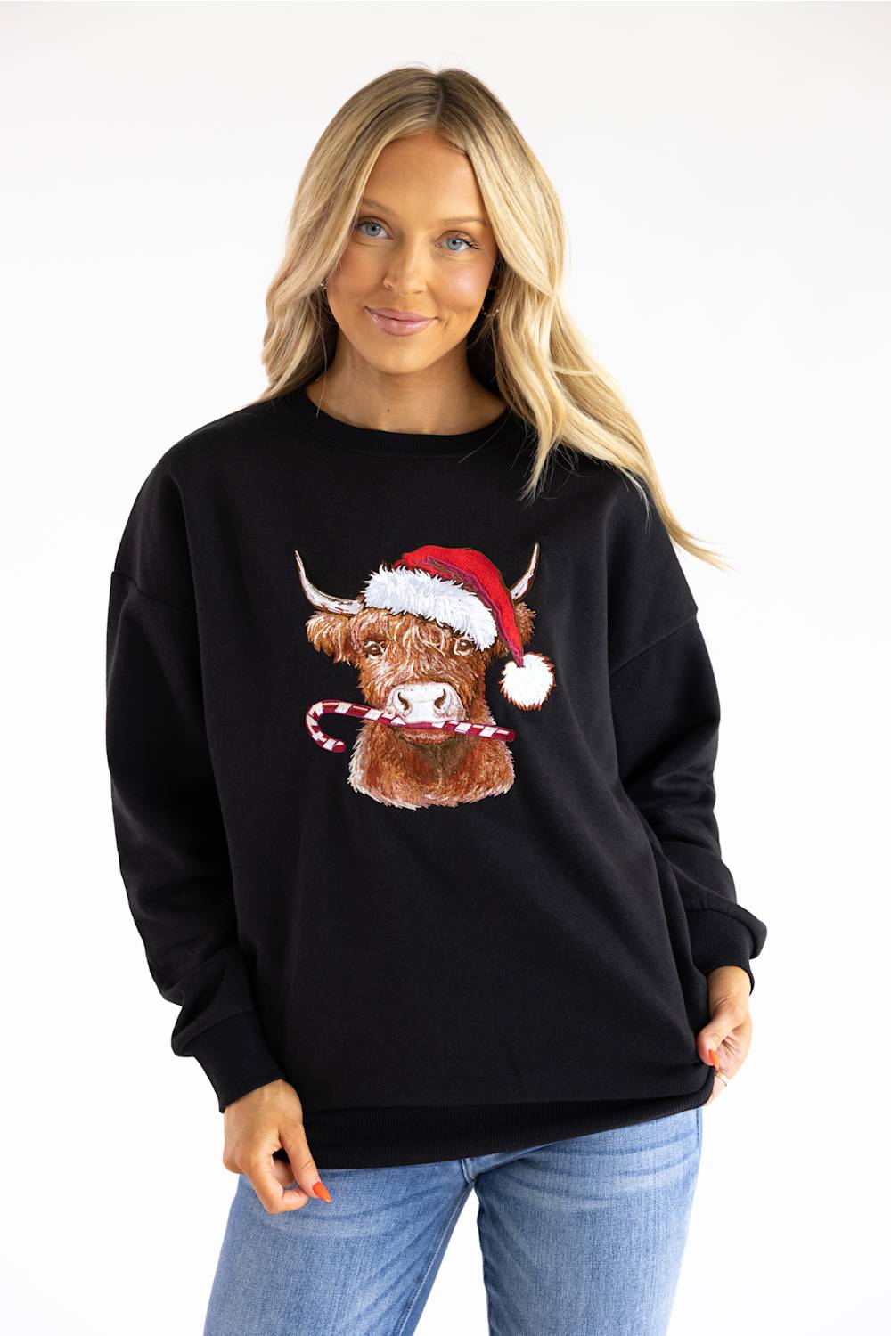 A woman wearing a black crewneck sweatshirt with a graphic of a Highland cow wearing a red Santa hat and holding a candy cane, paired with blue jeans, against a plain white background.