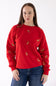 A woman wearing a bright red crewneck sweatshirt with small gold embroidered details, paired with blue jeans, against a plain white background.