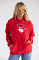 A woman wearing a bright red pullover hoodie with a festive goose graphic on the chest, paired with blue jeans, photographed against a light gray studio background.