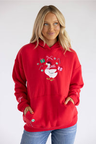 A woman wearing a bright red pullover hoodie with a festive goose graphic on the chest, paired with blue jeans, photographed against a light gray studio background.