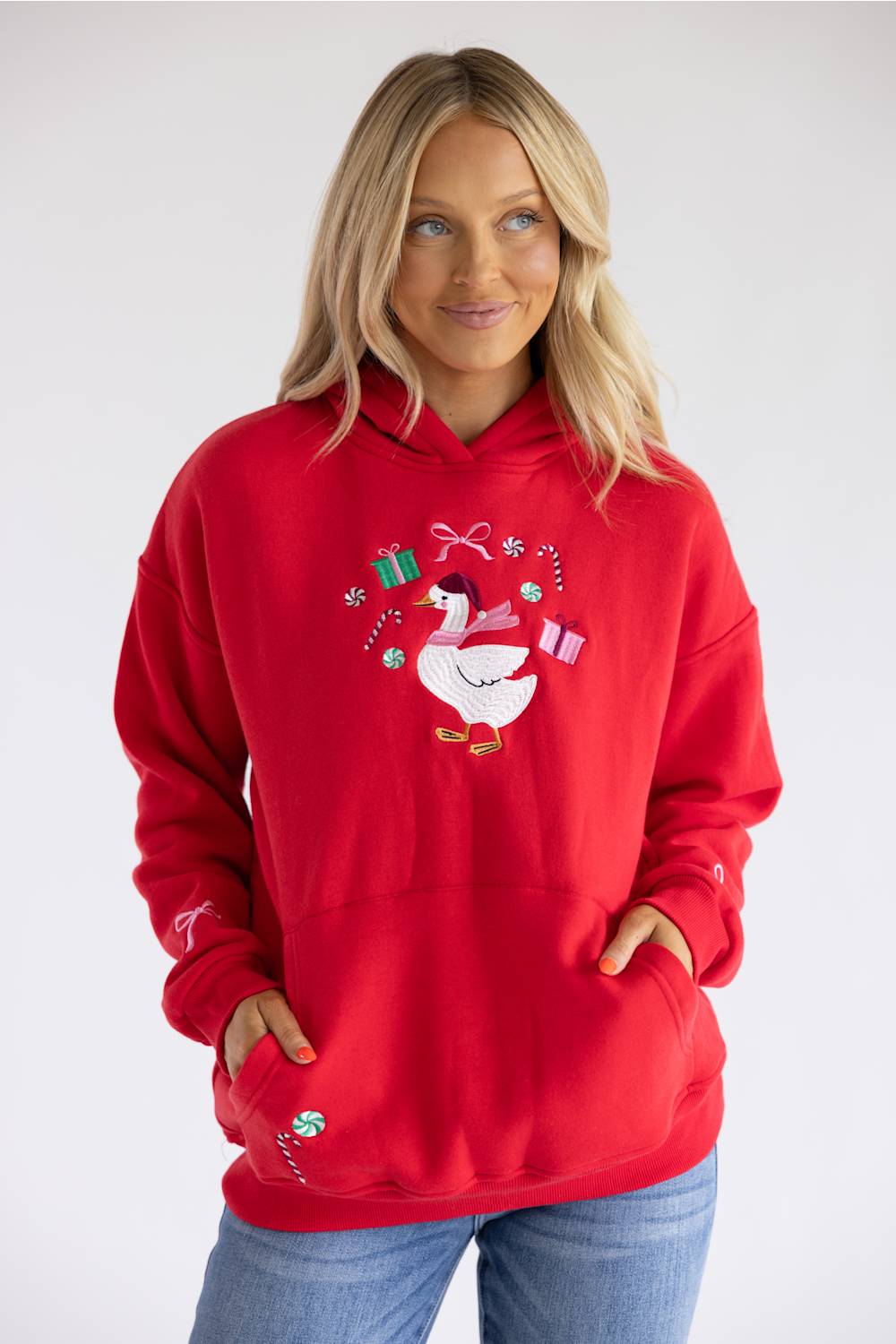 A woman wearing a bright red pullover hoodie with a festive goose graphic on the chest, paired with blue jeans, photographed against a light gray studio background.