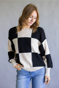 A woman wearing a black and cream checkered knit sweater with blue jeans, standing against a neutral gray background.