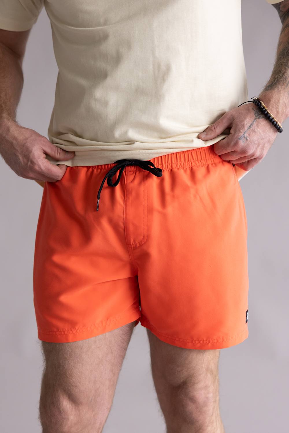 Front view of a person wearing orange bard shorts with a cream colored t-shirt on a neutral background. The shorts are the Quiksilver Everyday Solid Volley 15" Swim Shorts for Men in Hot Coral