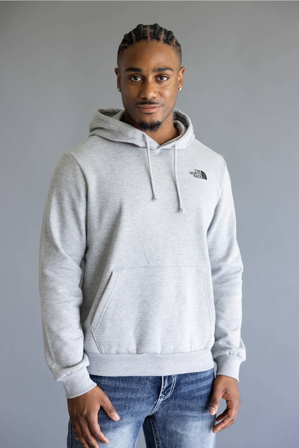 A man wearing a light gray heather pullover hoodie with a drawstring hood and kangaroo pocket, featuring the black embroidered logo of The North Face on the left chest, paired with blue jeans against a solid gray studio background.