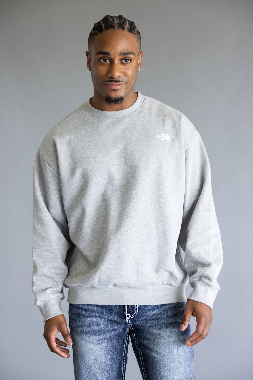 A man wearing a light gray crewneck sweatshirt and blue jeans, standing against a solid gray studio background.