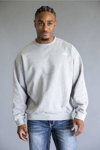 A man wearing a light gray crewneck sweatshirt and blue jeans, standing against a solid gray studio background.