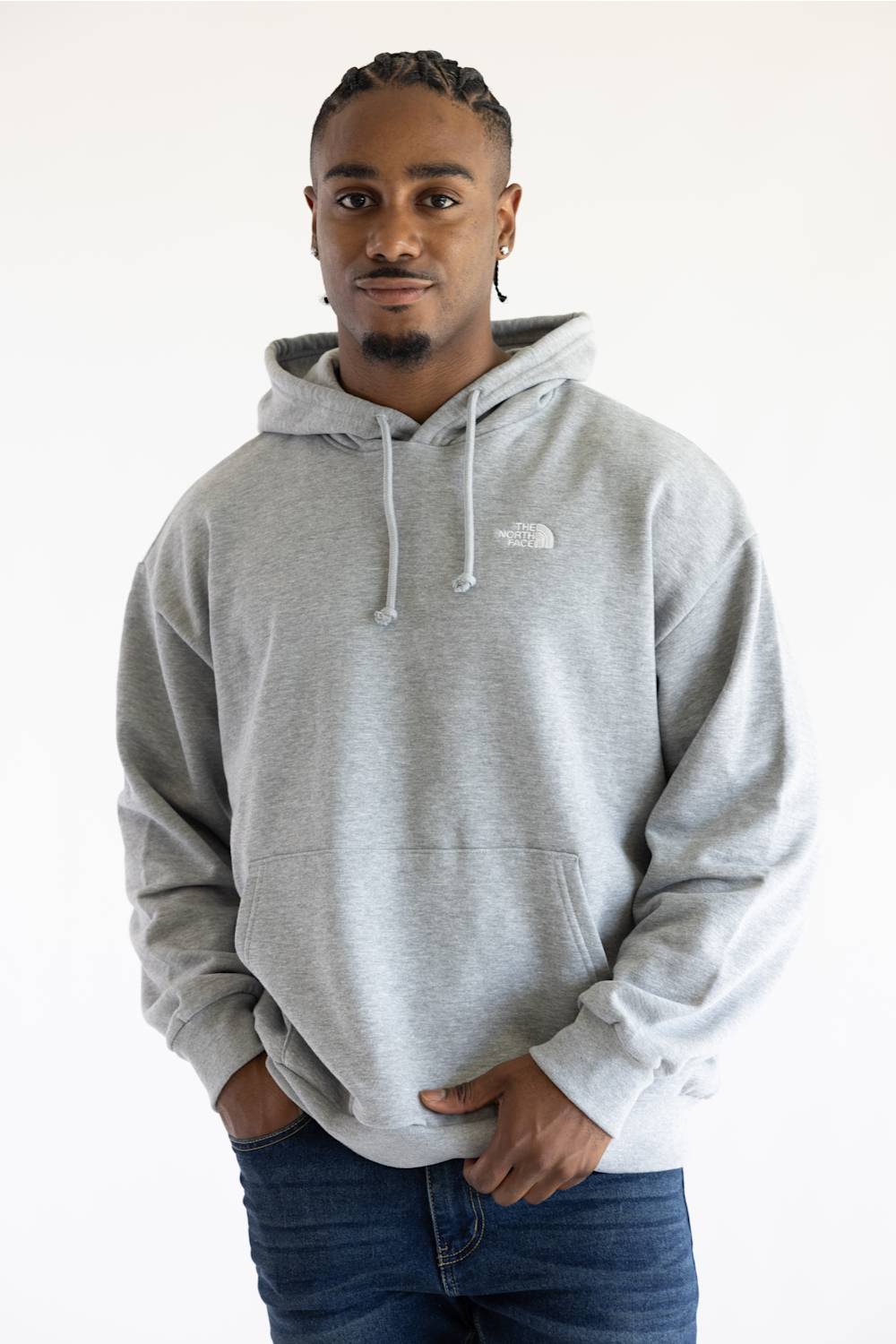 A man wearing a light gray heather pullover hoodie with a drawstring hood and kangaroo pocket, featuring the white embroidered logo of The North Face on the left chest, paired with blue jeans against a plain white background.