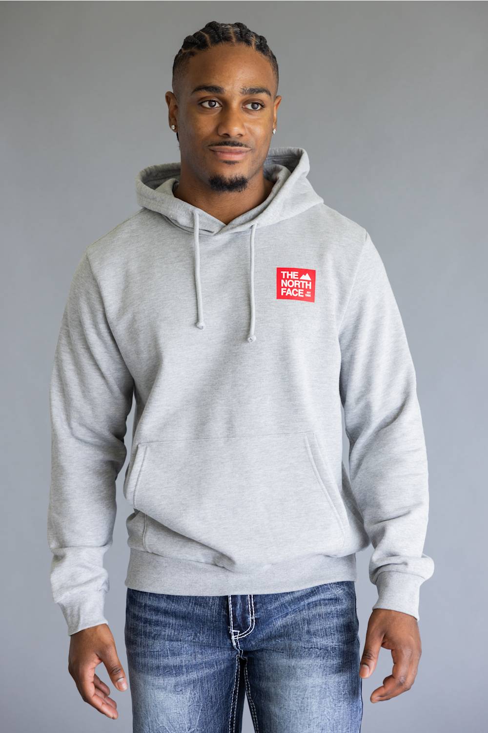 A man wearing a light gray heather pullover hoodie with a red 'The North Face' logo patch on the left chest, paired with blue jeans, against a solid gray studio background.