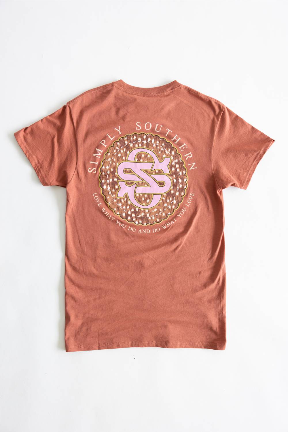 A terracotta-colored short-sleeve t-shirt laid flat against a white background, featuring a large circular graphic on the back with the text 'SIMPLY SOUTHERN' and a pink-and-gold monogram logo.