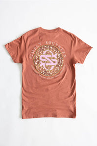 A terracotta-colored short-sleeve t-shirt laid flat against a white background, featuring a large circular graphic on the back with the text 'SIMPLY SOUTHERN' and a pink-and-gold monogram logo.