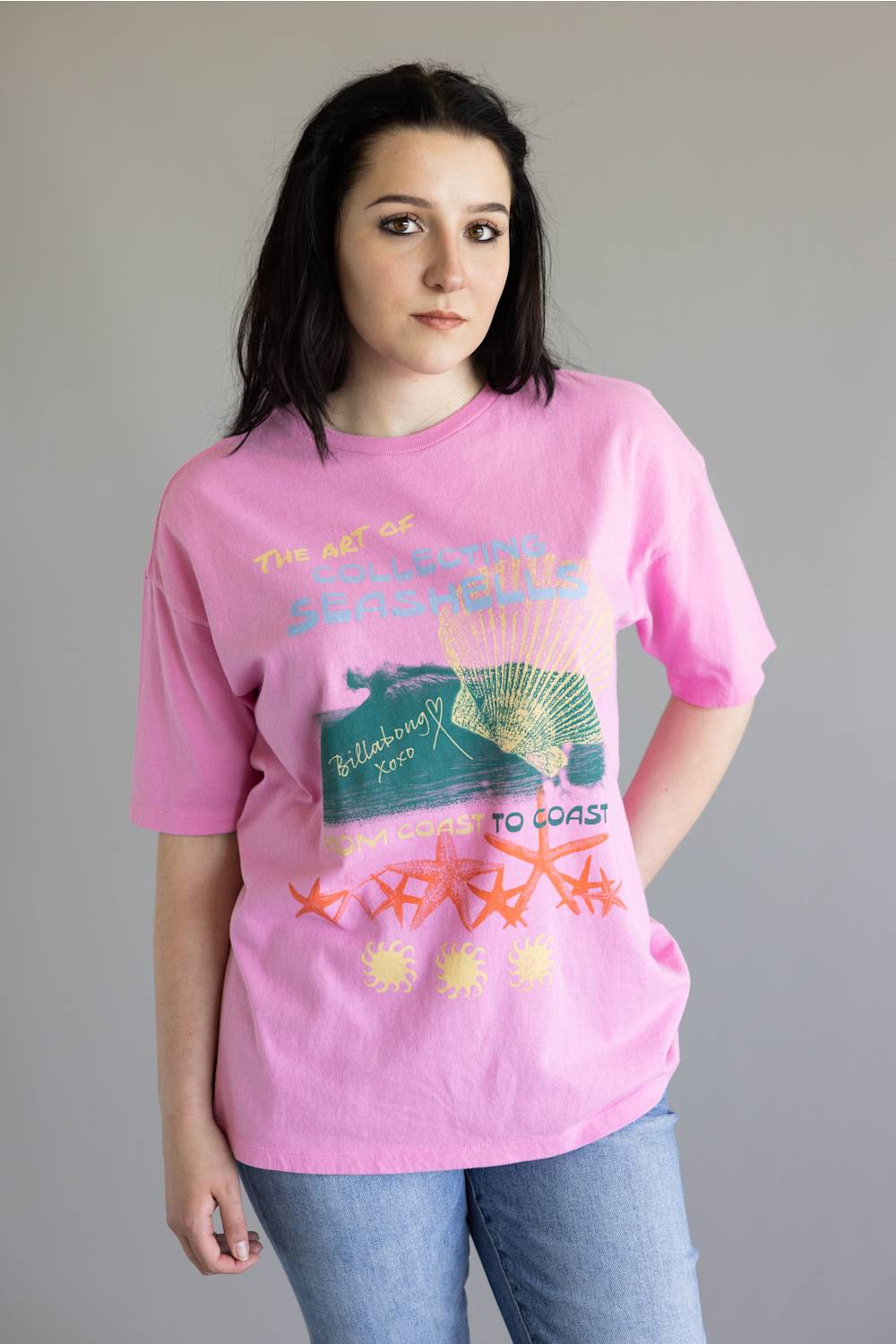 A woman wearing a pink graphic t-shirt with a vintage-style design featuring starfish and text, paired with blue jeans, photographed against a plain gray background.