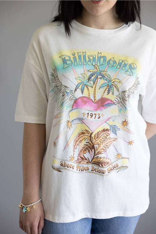 A person wearing a white Billabong graphic t-shirt with a colorful tropical print featuring palm trees, a pink heart, and the text '1973' and 'Above Tide Below Tide'.