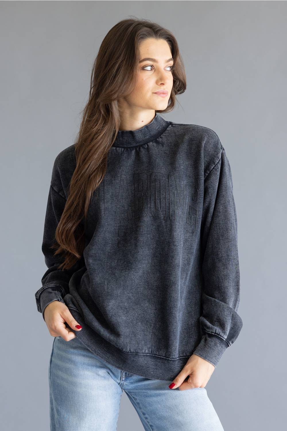 A woman wearing a dark gray, washed cotton crewneck sweatshirt and light blue jeans, standing against a solid gray studio background.