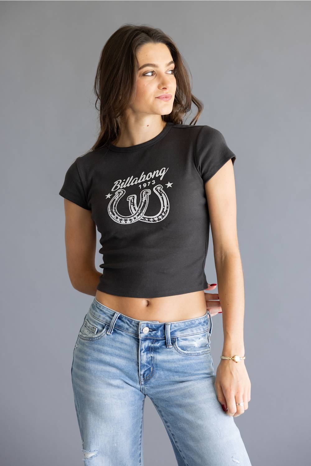 A woman modeling a black short-sleeve crop top with a white Billabong logo graphic on the chest, paired with light blue jeans, against a solid gray studio background.