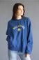 A woman wearing a blue crewneck sweatshirt with a yellow Billabong sun logo and text on the chest, paired with light wash jeans, against a solid gray studio background.