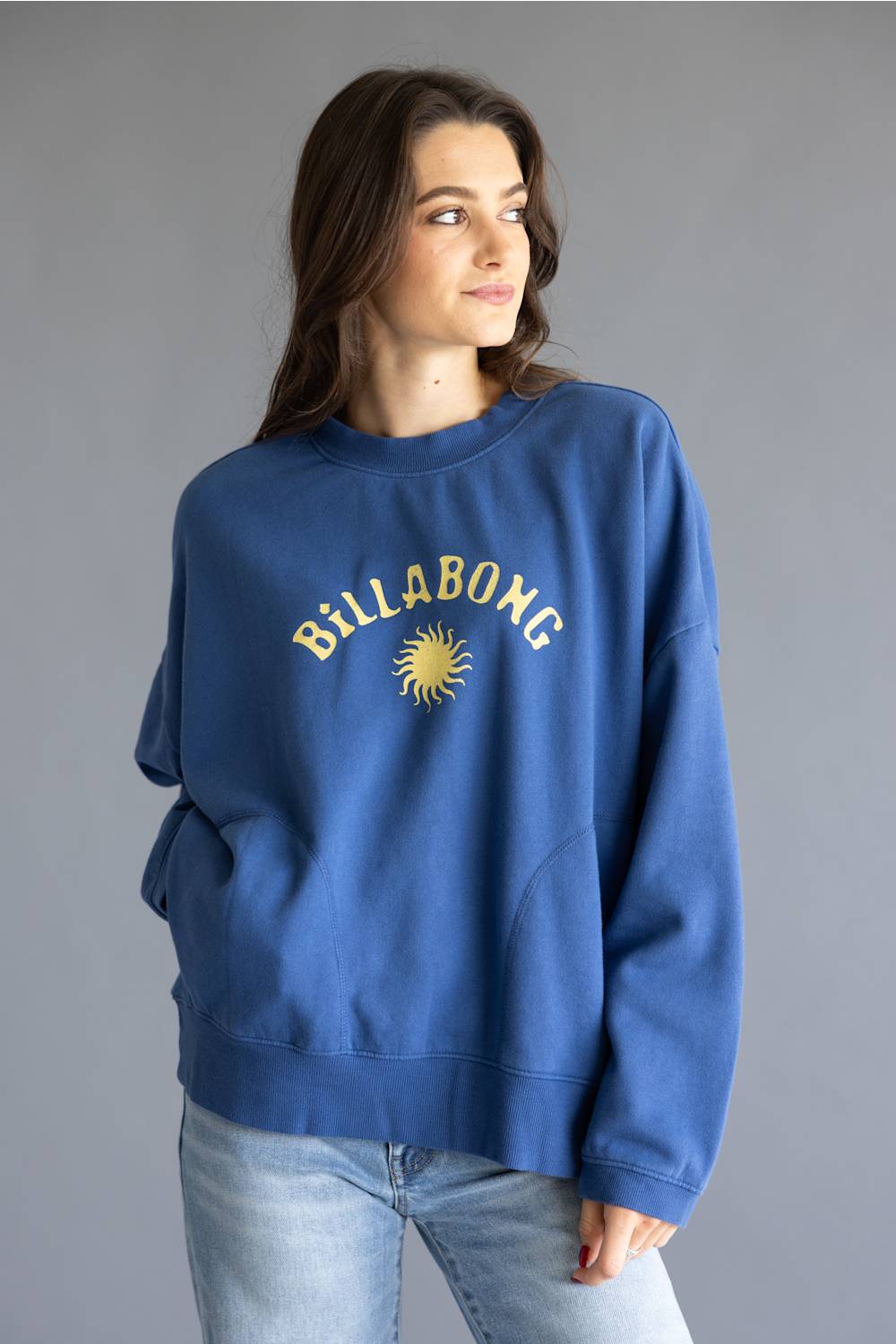 A woman wearing a blue crewneck sweatshirt with a yellow Billabong sun logo and text on the chest, paired with light wash jeans, against a solid gray studio background.