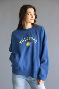 A woman wearing a blue crewneck sweatshirt with a yellow Billabong sun logo and text on the chest, paired with light wash jeans, against a solid gray studio background.