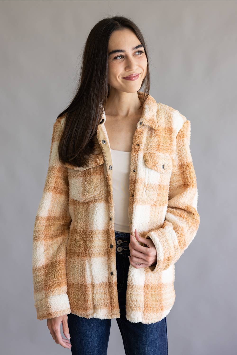 A woman wearing a fuzzy plaid button-up jacket in cream and tan tones, paired with a white top and dark blue jeans, standing against a neutral gray background.