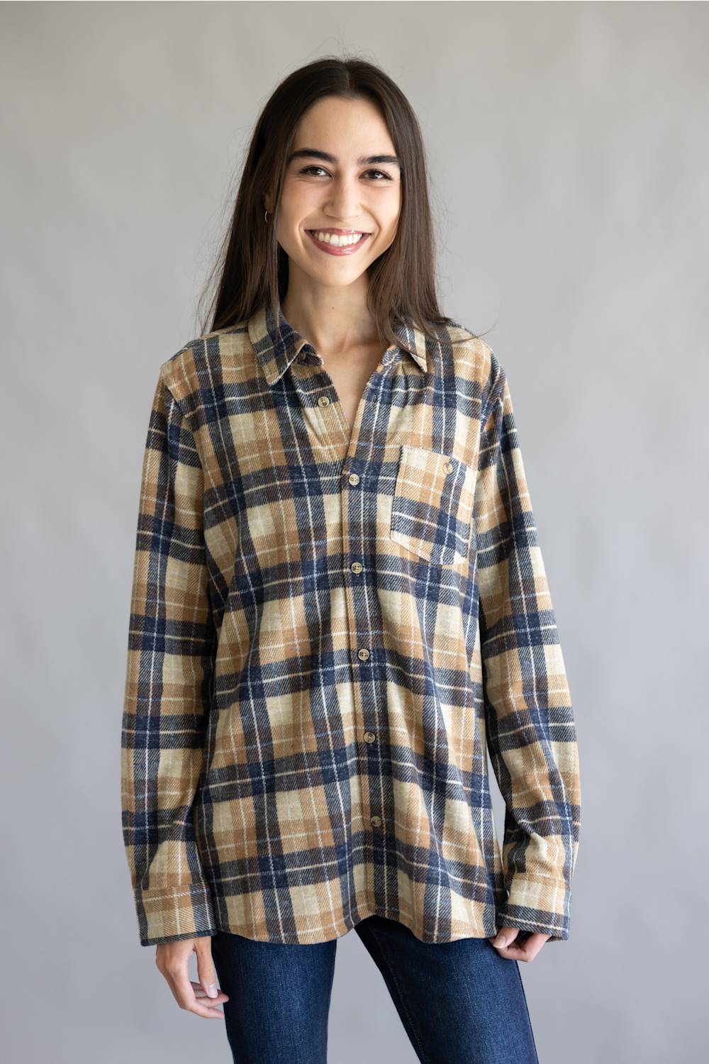 A woman wearing a long-sleeve button-up plaid shirt in beige, navy, and brown tones, paired with dark blue jeans, standing against a plain gray studio background.