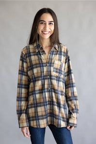 A woman wearing a long-sleeve button-up plaid shirt in beige, navy, and brown tones, paired with dark blue jeans, standing against a plain gray studio background.