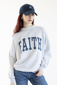 A woman wearing a light heather gray crewneck sweatshirt with the word 'FAITH' printed across the chest in navy blue block letters filled with white polka dots, paired with blue jeans.