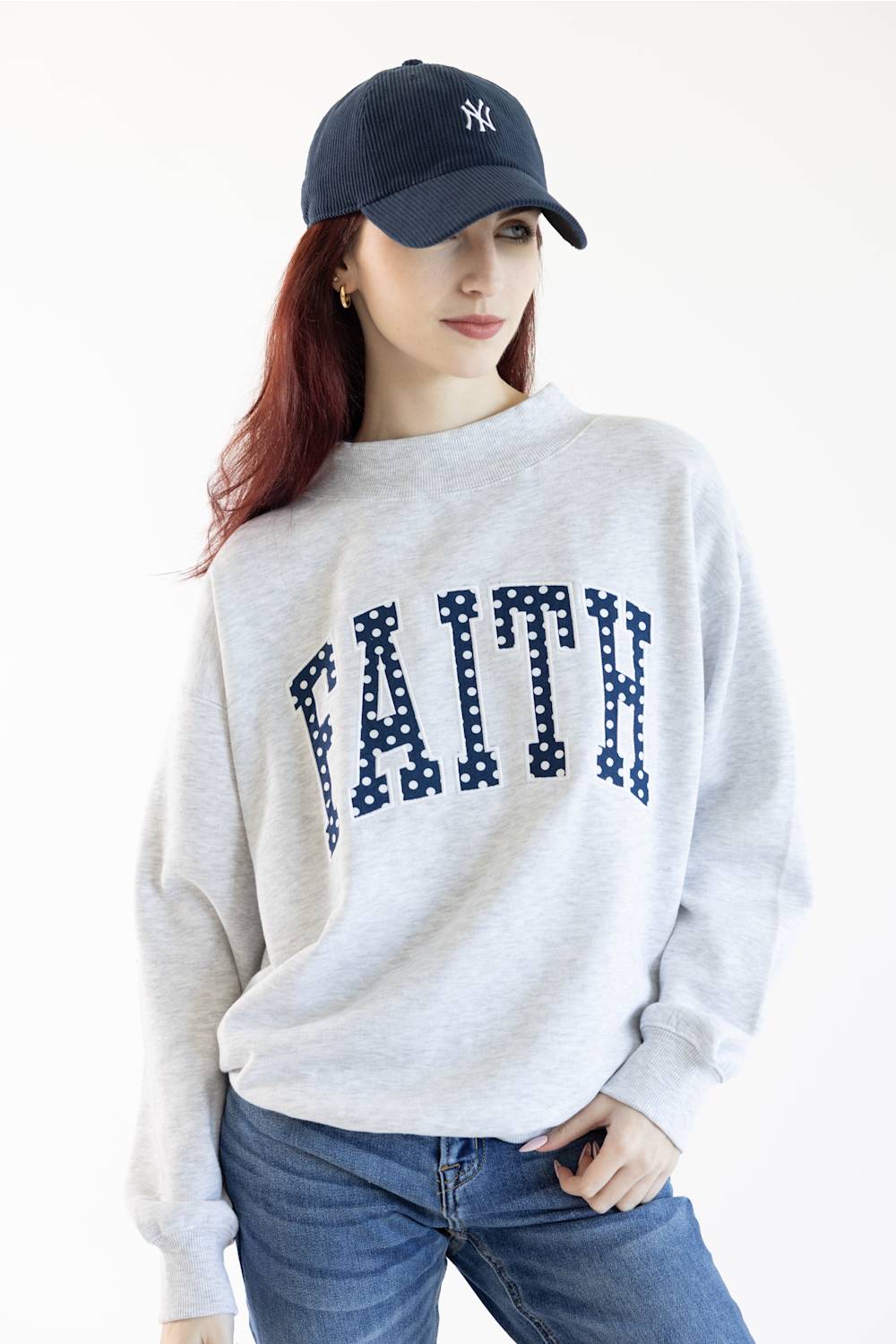 A woman wearing a light heather gray crewneck sweatshirt with the word 'FAITH' printed across the chest in navy blue block letters filled with white polka dots, paired with blue jeans.